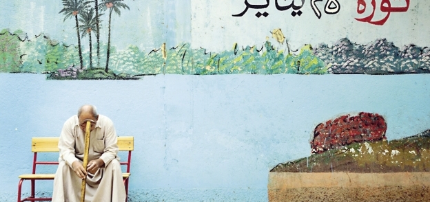 مواطن يجلس أمام لجنة انتخابية بإحدى المدارس وخلفه جرافيتى عن ثورة يناير
